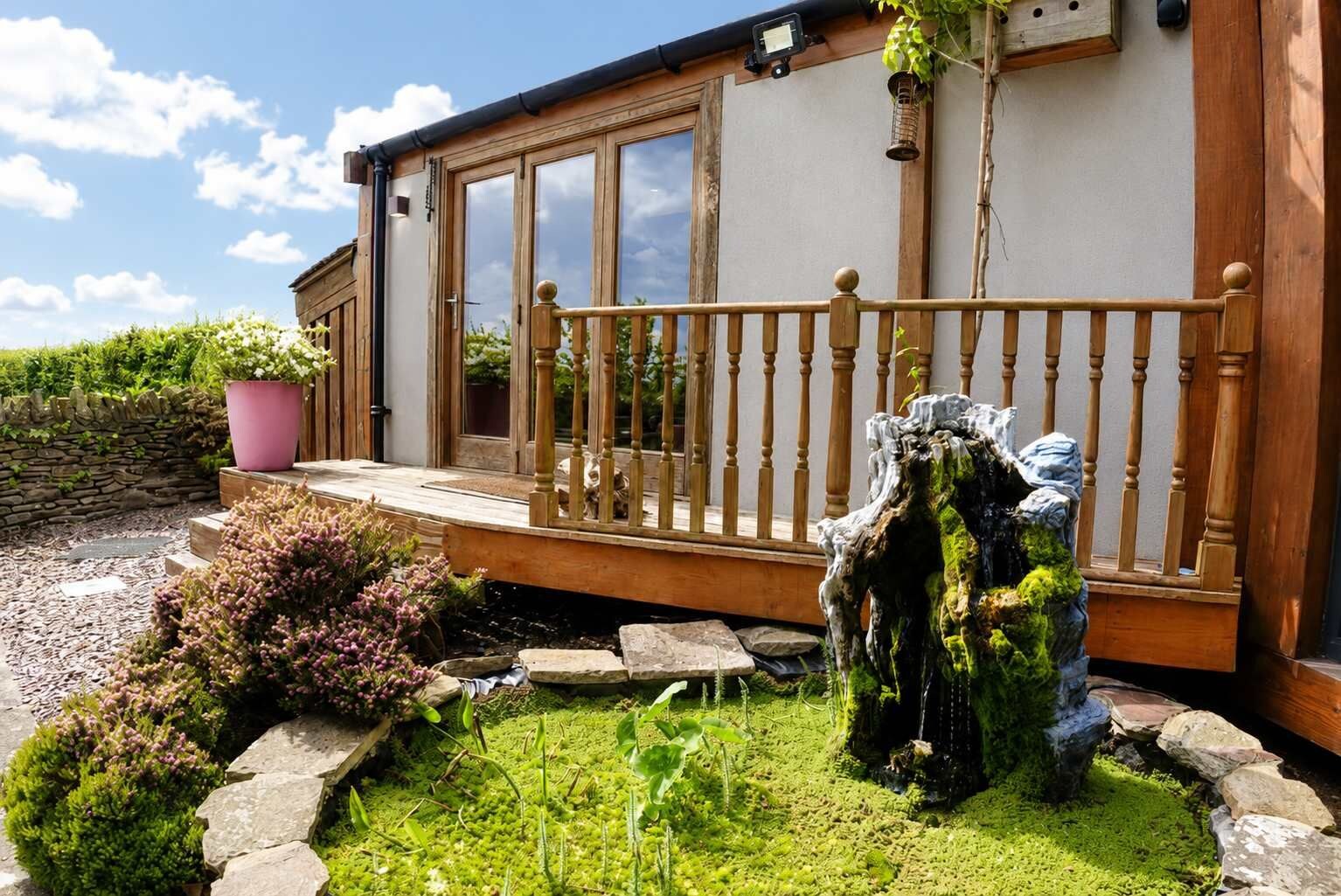 A wooden house with a balcony, a stone wall, and a garden with plants and a rock feature.