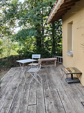 A wooden deck with a table and chairs, surrounded by greenery and a building. - Studio in the countryside with a view of the Pyrenees. (Castillon-de-Saint-Martory)