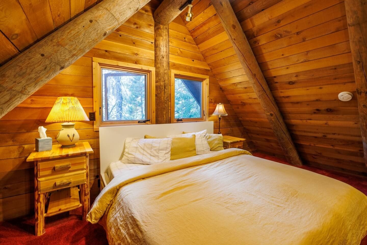 A cozy wooden cabin bedroom with a bed, bedside lamps, and a window providing a view of trees.