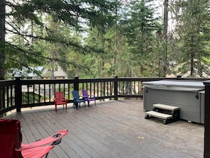 Een houten terras met een zwarte balustrade, omringd door bomen, met een jacuzzi en buitenmeubilair.