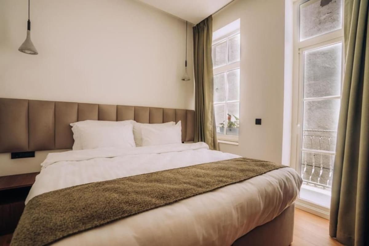 A hotel room with a bed, a bedside table, a window with curtains, and a view of the outside.
