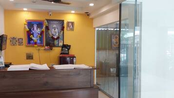 A reception desk with a wooden finish, a glass partition, and a yellow wall with religious posters and a framed picture.