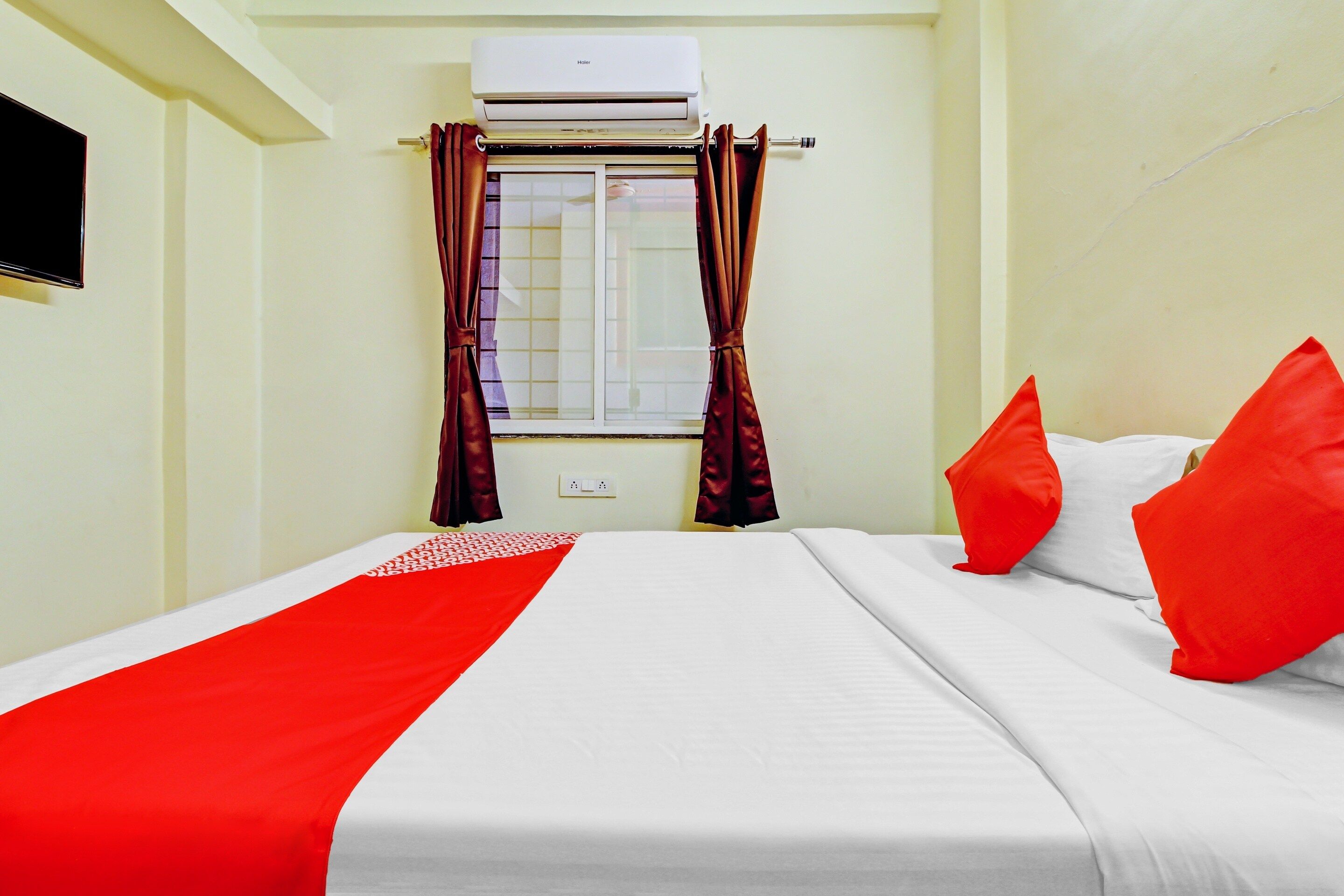 Una habitación de hotel con cama, almohadas rojas, colcha roja y blanca, ventana con cortinas y un equipo de aire acondicionado.