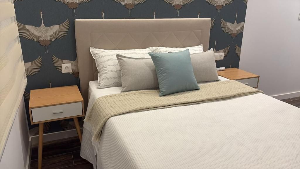 Una cama bien hecha con cabecera, dos mesitas de noche y una pared con un motivo de pájaros.