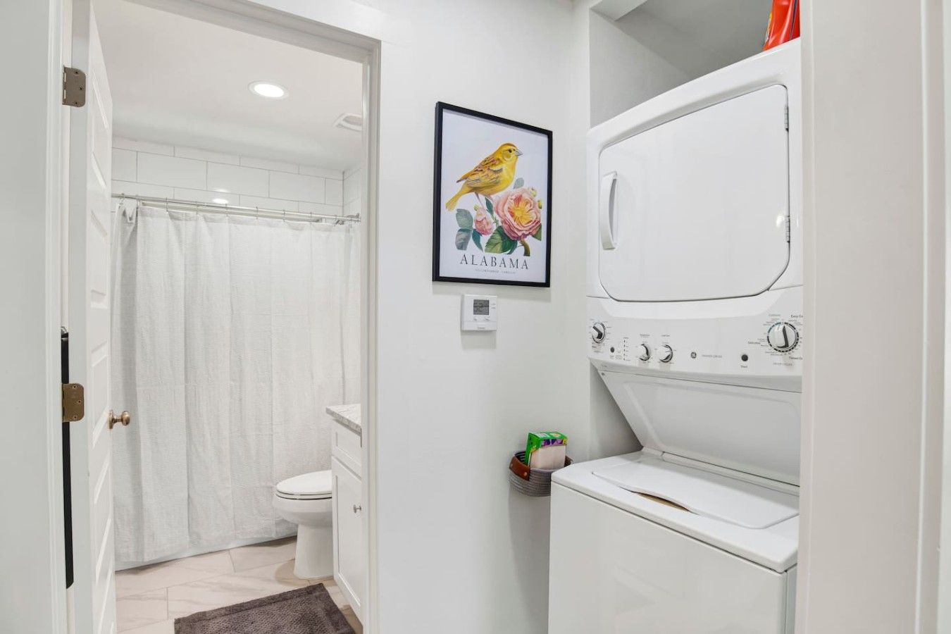 A compact bathroom with a toilet, a shower curtain, a framed picture of a bird, and a white washing machine.