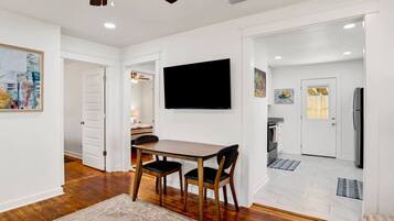 A modern interior with a dining area, a flat-screen TV mounted on the wall, and a kitchen visible through an open door.