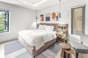 Habitación de hotel moderna con una cama grande, una mesita de noche con teléfono, una silla y una ventana con vistas a zonas verdes.