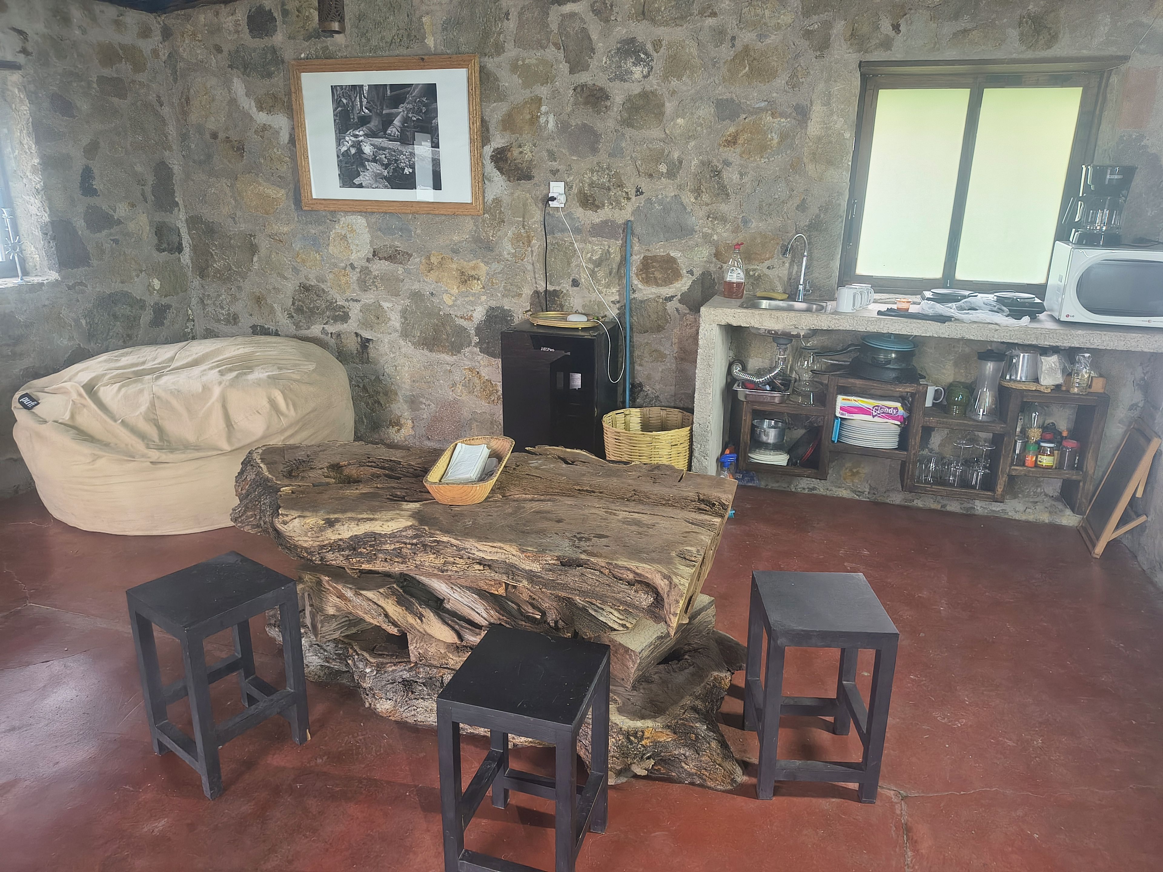 Un interior rústico con una pared de piedra, una fotografía enmarcada, una mesa de madera con taburetes y una zona de kitchenette.
