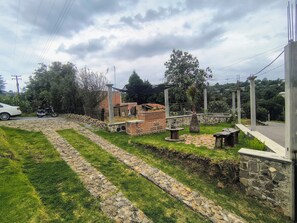 A paved pathway leading to a brick structure with a small table and a palm tree. - Cabaña Tetitla — Una escapada entre naturaleza y tradición en Tlaxcala. (Amaxac de Guerrero)