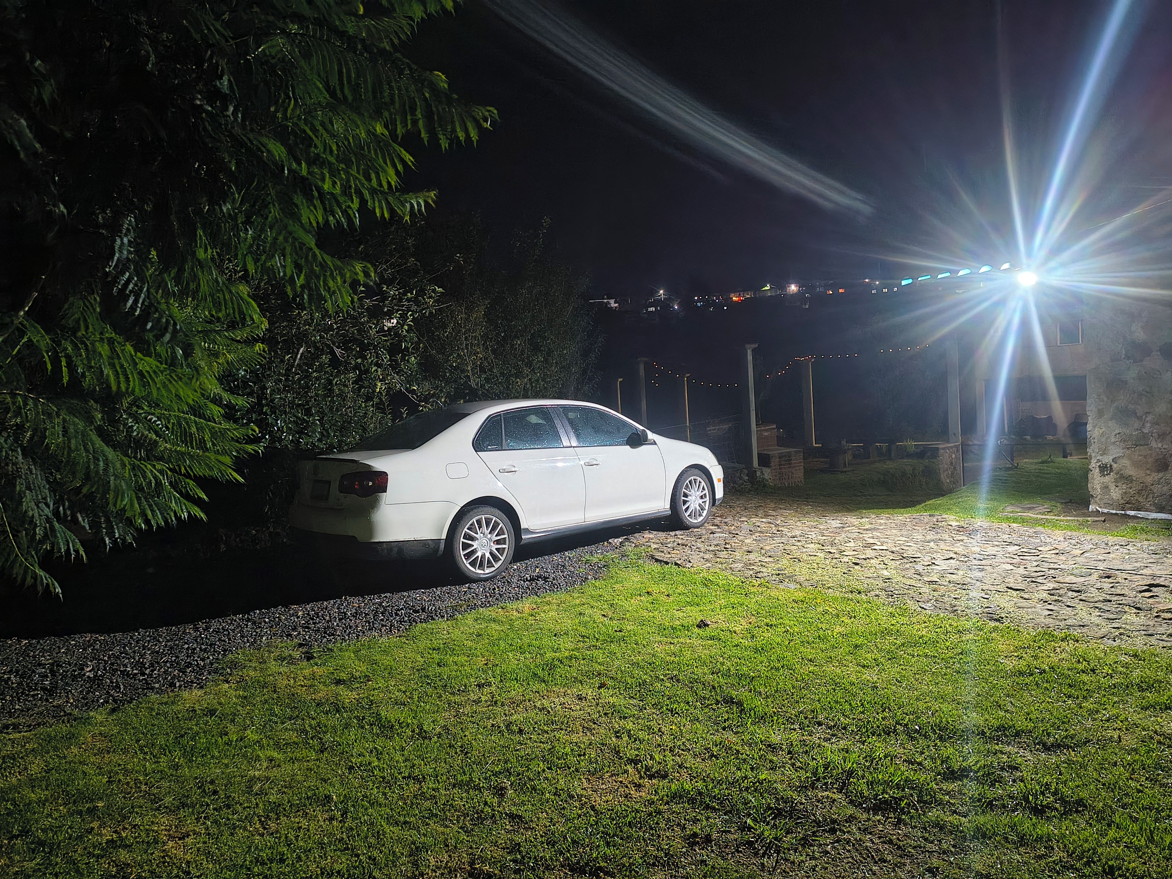 Un coche blanco aparcado en un camino de grava por la noche, con una farola al fondo.