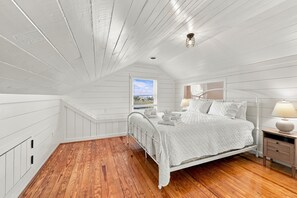 A bedroom with a sloped ceiling, a bed with white bedding, a nightstand with a lamp, and a window with a view. - Marsh Bend: 8  BR, 5  BA House in Pawleys Island, Sleeps 21 (Pawleys Island)