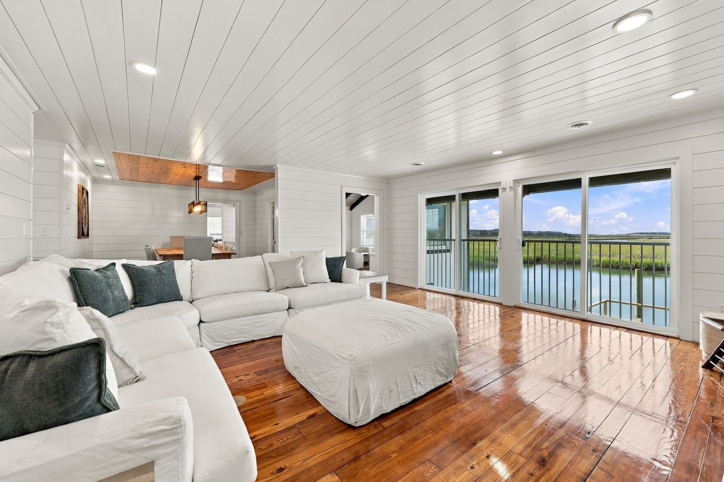 A spacious living room with a large sectional sofa, a small ottoman, and a wooden floor. There are large windows offering a view of a body of water and greenery.
