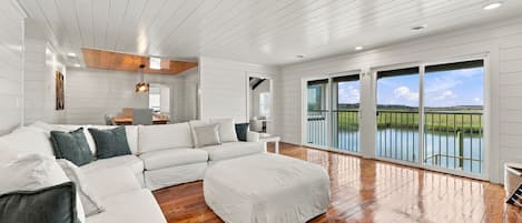 A spacious living room with a large sectional sofa, a small ottoman, and a wooden floor. There are large windows offering a view of a body of water and greenery.
