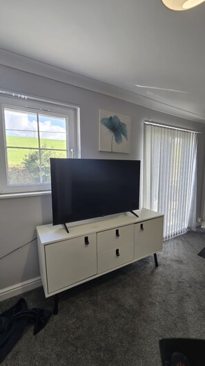 Habitació amb televisió de pantalla plana sobre un moble blanc, una finestra amb vista a un camp verd i un llum al sostre.