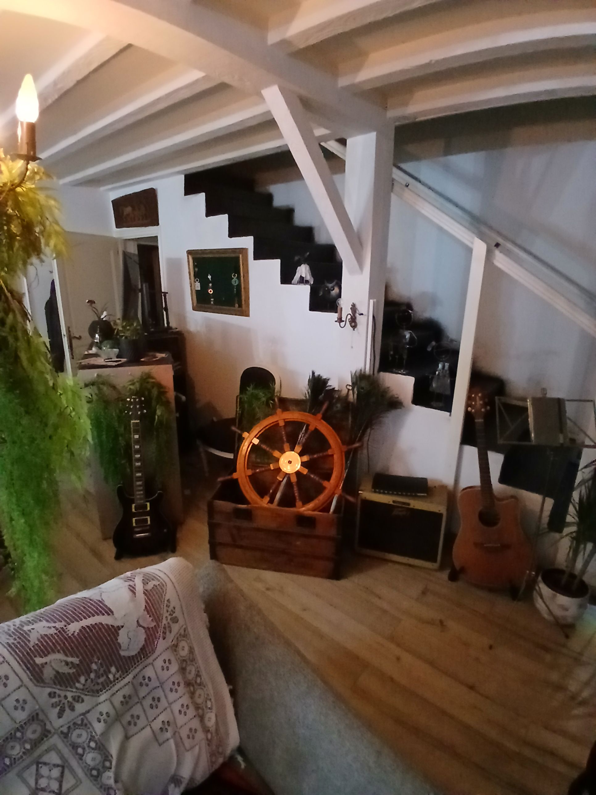 Una stanza accogliente con pavimento in legno, una poltrona a dondolo, un tavolino con una pianta, un orologio a muro e una scala che conduce a un piano superiore.