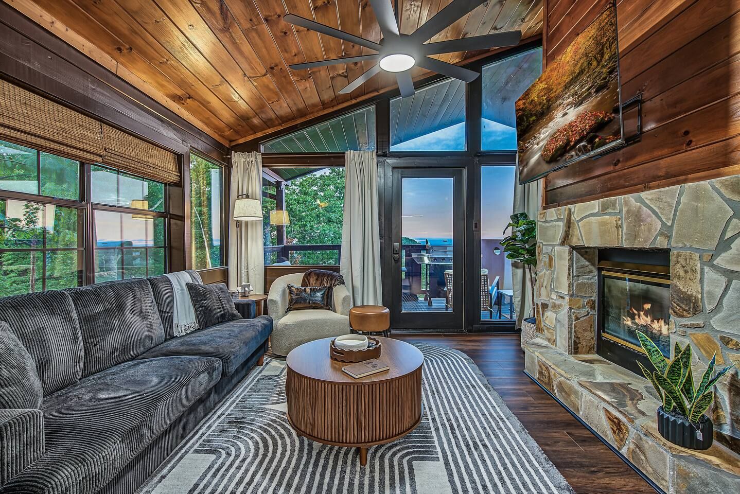 A cozy living room with a fireplace, a sofa, a coffee table, and a view of the outdoors through large windows.