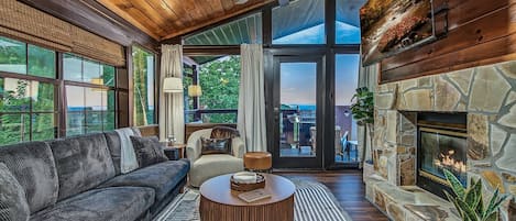 A cozy living room with a fireplace, a sofa, a coffee table, and a view of the outdoors through large windows.