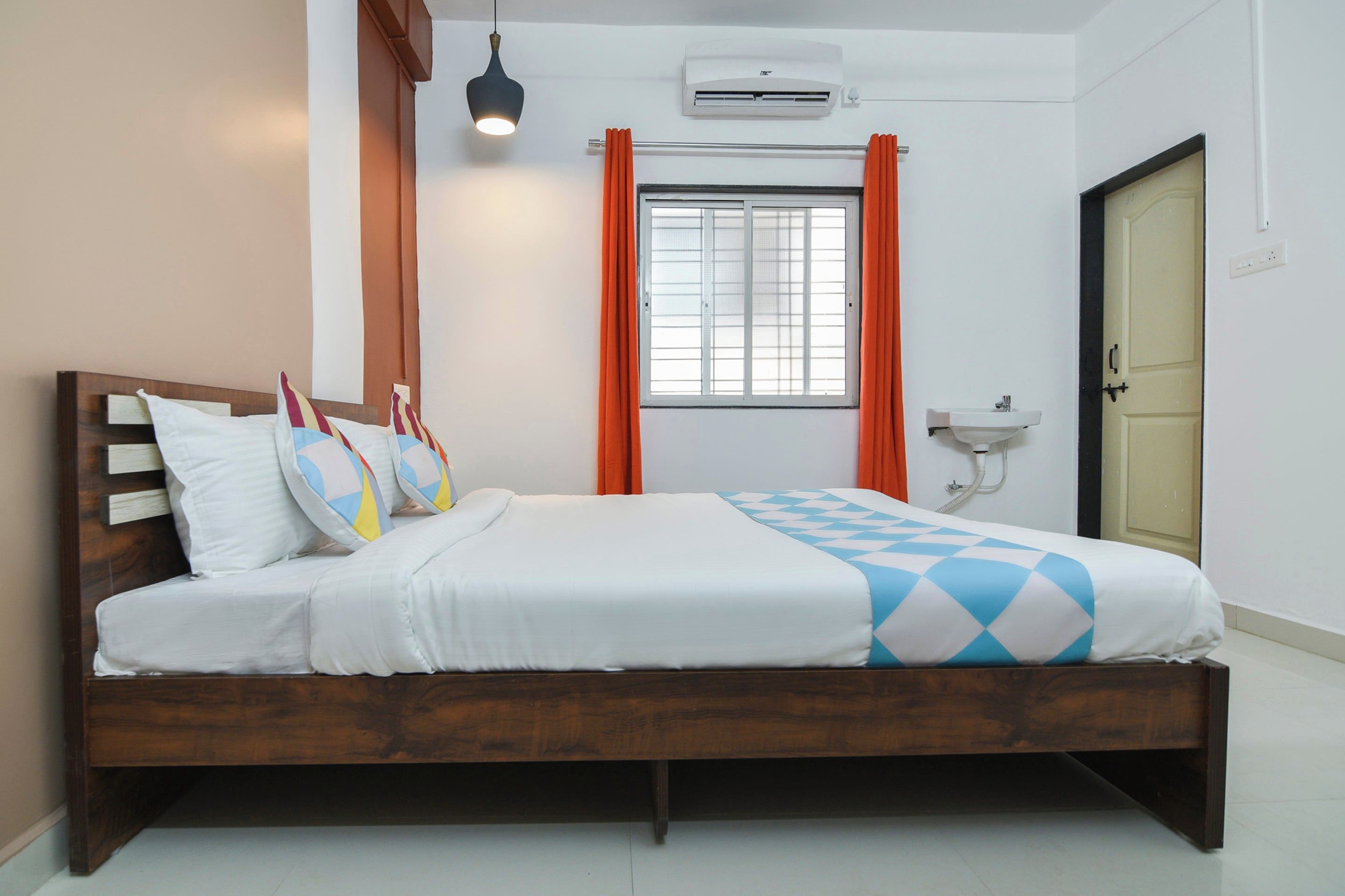 A hotel room with a wooden bed, a window with orange curtains, an air conditioning unit, and a bedside table with a sink.