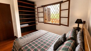 A bedroom with a bed, a window with blinds, a wooden wardrobe, and a bedside table with a lamp.