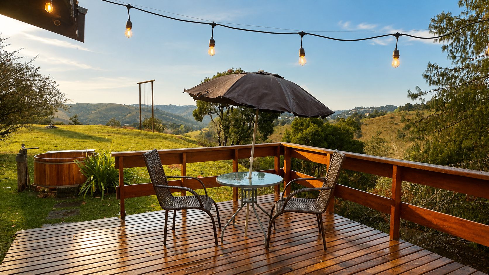Um deck de madeira com mesa e cadeiras, cercado por um corrimão, com vista para uma paisagem montanhosa.