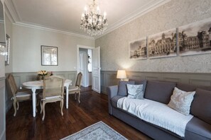 Ein Wohnzimmer mit dunklem Holzboden, einem grauen Sofa, einer Chandelier und einem Essbereich mit weißem Tisch und Korbstühlen.