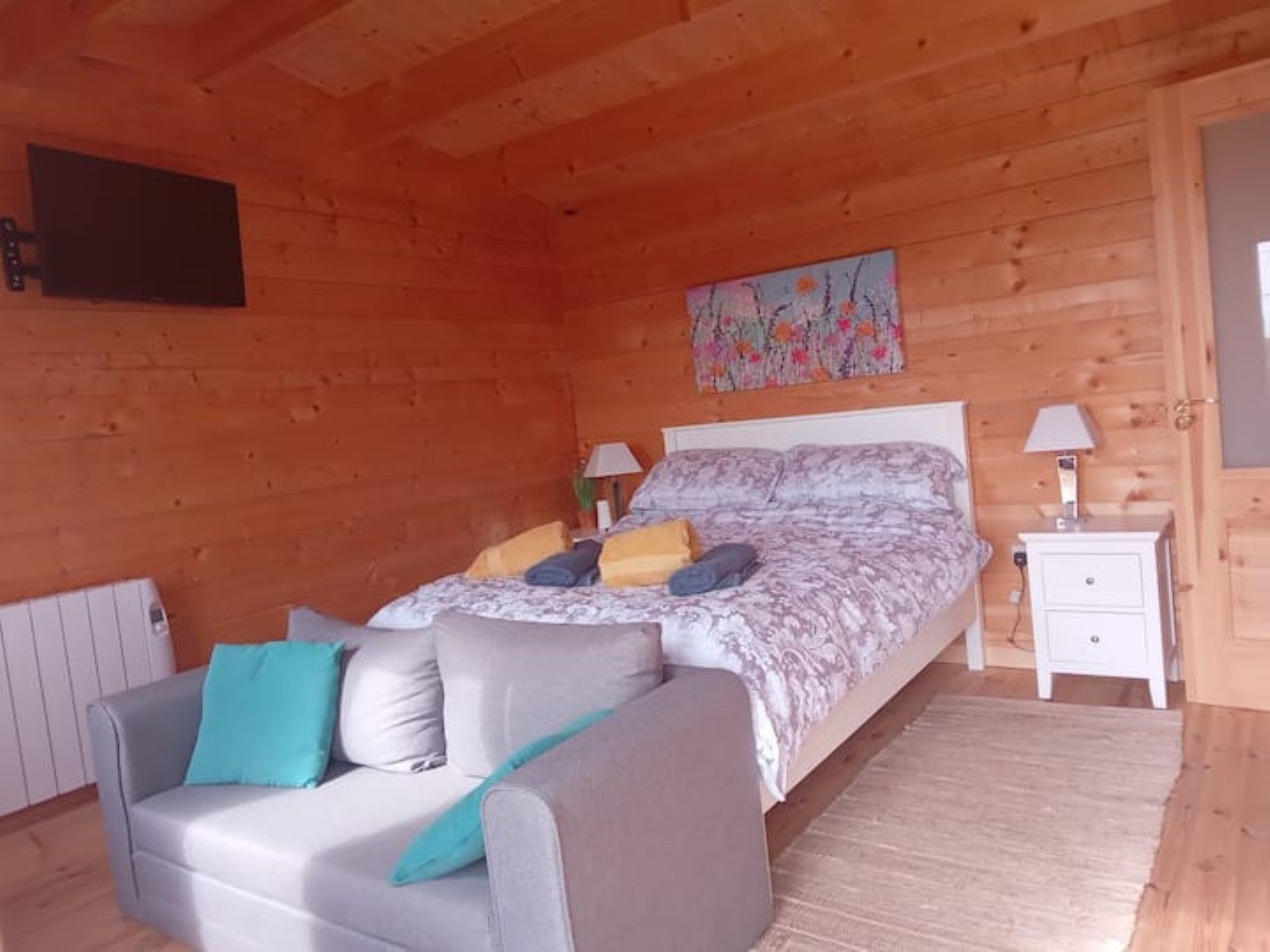 A wooden cabin bedroom with a bed, a sofa, a nightstand, and a radiator.