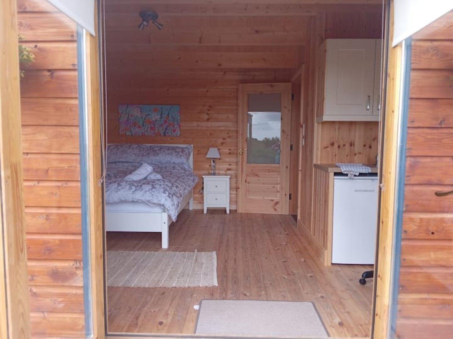 Una pequeña cabaña de madera con una cama individual, una mesita de noche, un escritorio y un refrigerador.