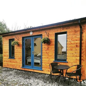 A wooden cabin with a blue door, two windows with hanging flower baskets, and a small outdoor seating area with two chairs. - COSY CUTE COUNTRYSIDE CABIN (CLARE)