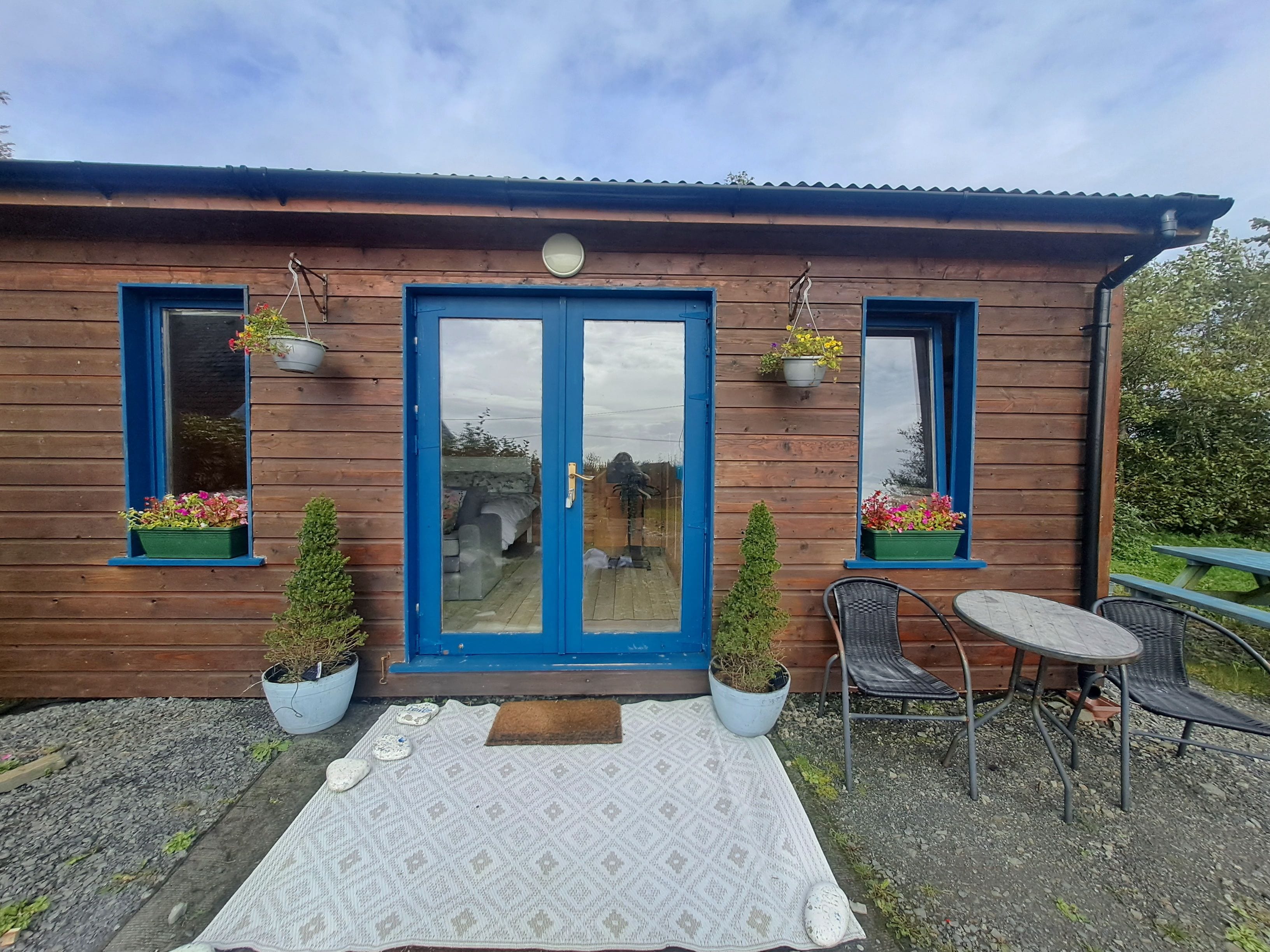 A wooden cabin with a blue door, two windows with hanging flower baskets, a small patio with a table and chairs, and a garden visible through the door.