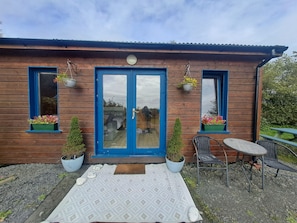 A wooden cabin with a blue door, two windows with hanging flower baskets, a small patio with a table and chairs, and a garden visible through the door. - COSY CUTE COUNTRYSIDE CABIN (CLARE)