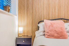 Quarto com parede de painel de madeira, mesa de cabeceira com abajur e cama com travesseiro rosa.