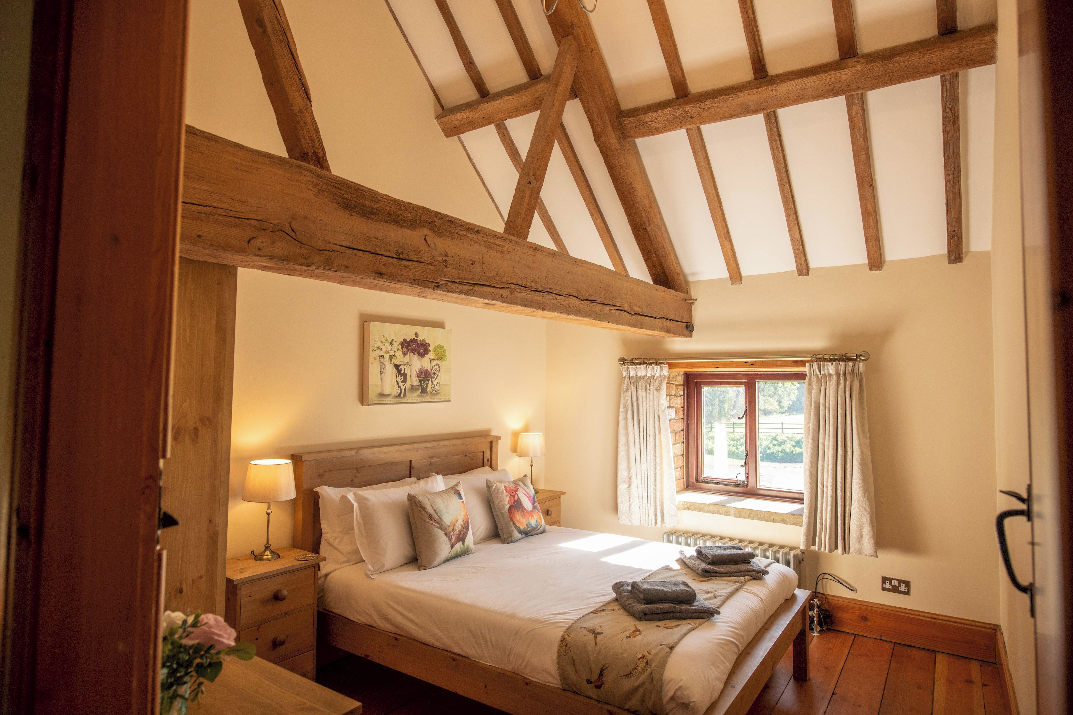 A bedroom with a wooden ceiling, a bed with multiple pillows, a nightstand with a lamp, and a window with curtains.