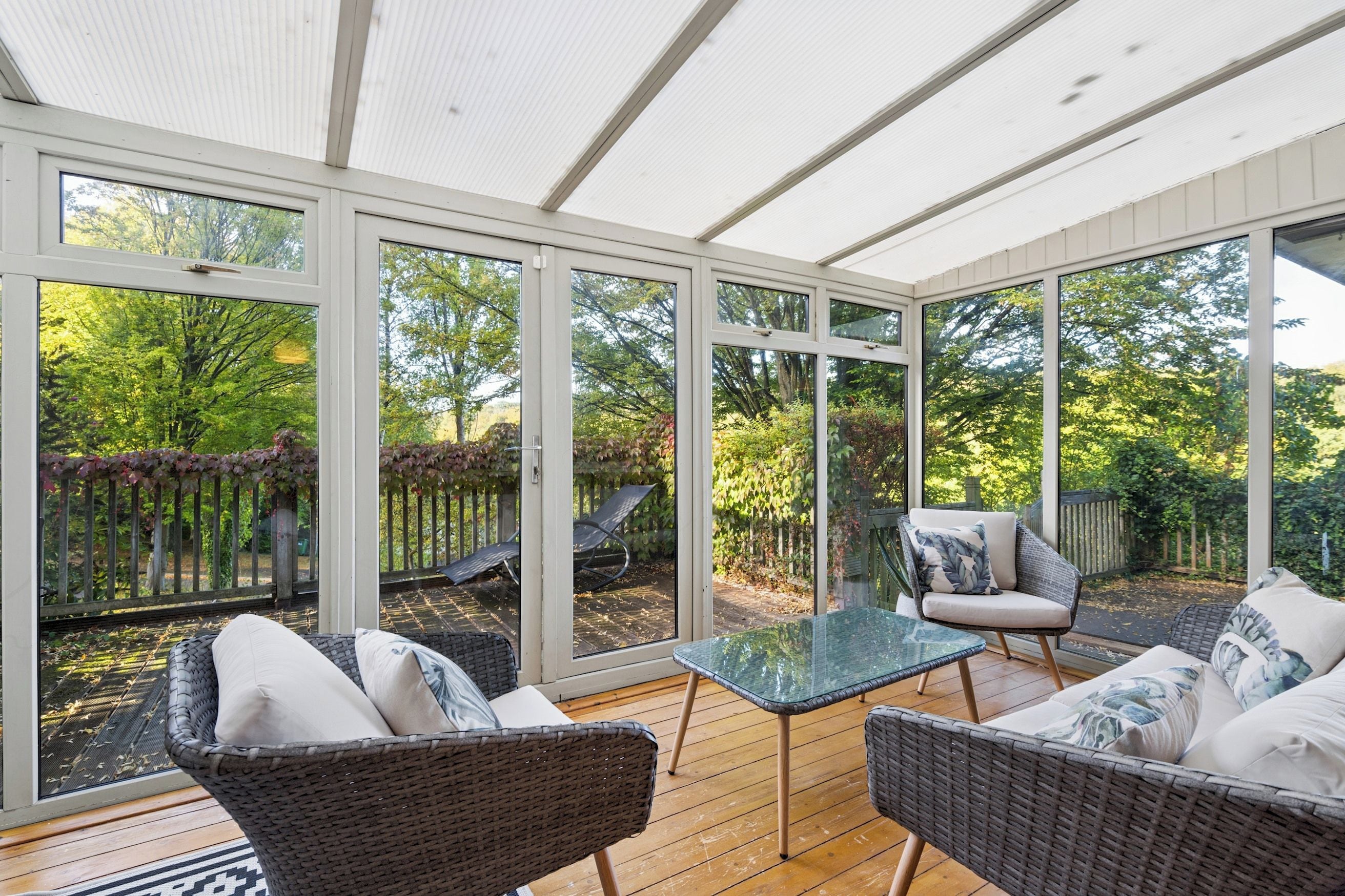 A conservatory with wicker furniture, glass table, and a view of a garden.