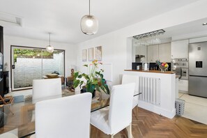 A modern kitchen with stainless steel appliances, a dining area with white chairs, and a large window overlooking a brick wall. - Chilterns Leafy Hillside Hideaway - Pass the Keys (High Wycombe)