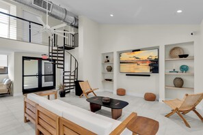 A modern living room with a sofa, coffee table, and chairs, a staircase, and a large TV displaying a sunset view. - Islamorada Townhouse - Sunsets, Beach & Sparkling Pool! (Islamorada)