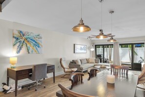A spacious living area with a dining table, sofas, and a desk. There are pendant lights, a painting, and a view through the glass door. - Islamorada Townhouse - Sunsets, Beach & Sparkling Pool! (Islamorada)