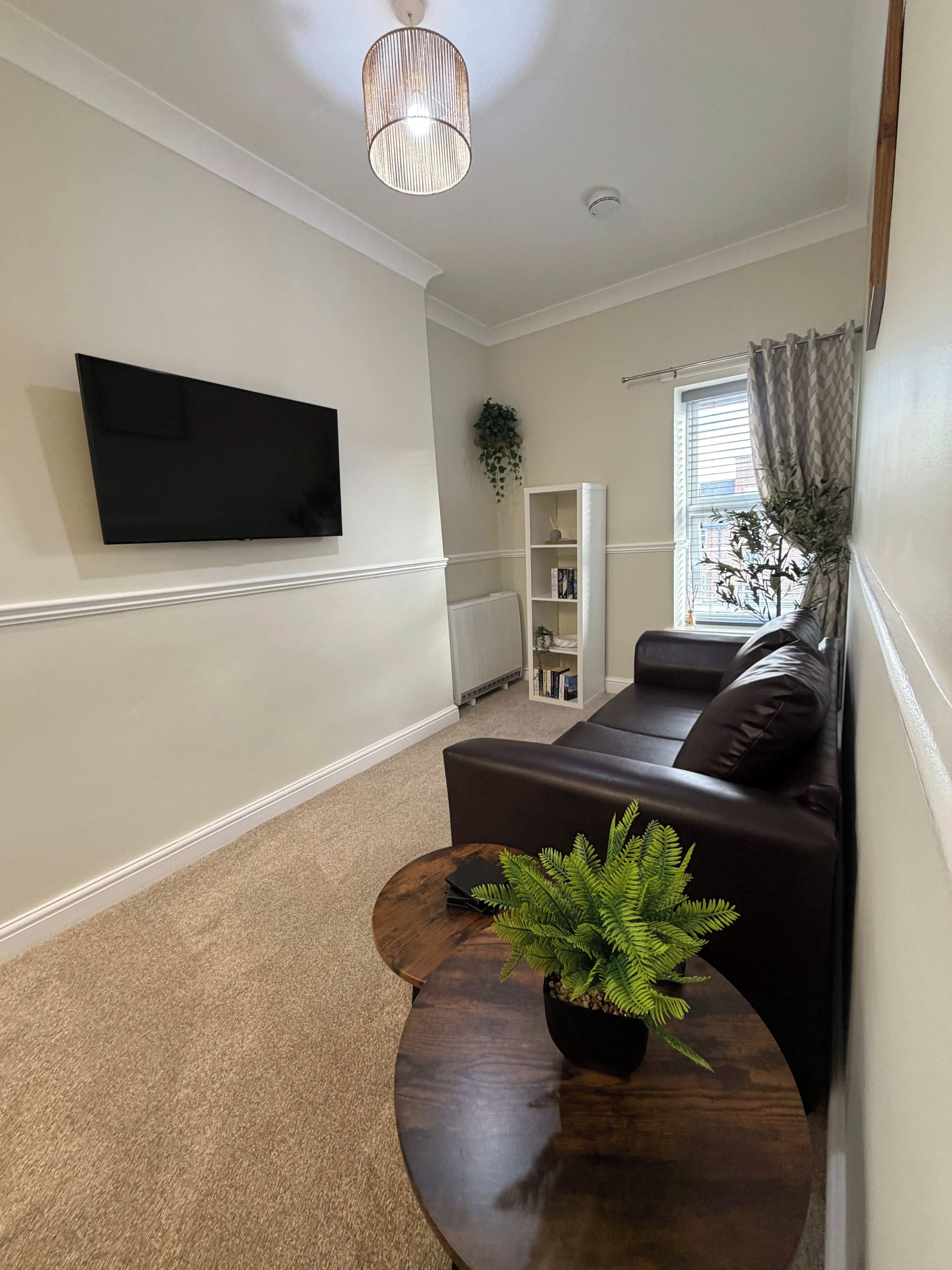 Een woonkamer met een flatscreen televisie aan de muur, een donkere bank, een ronde salontafel met een plant en een witte boekenkast.