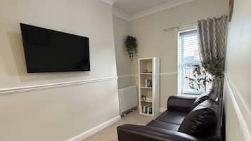 Een woonkamer met een flatscreen televisie aan de muur, een donkere bank, een ronde salontafel met een plant en een witte boekenkast.