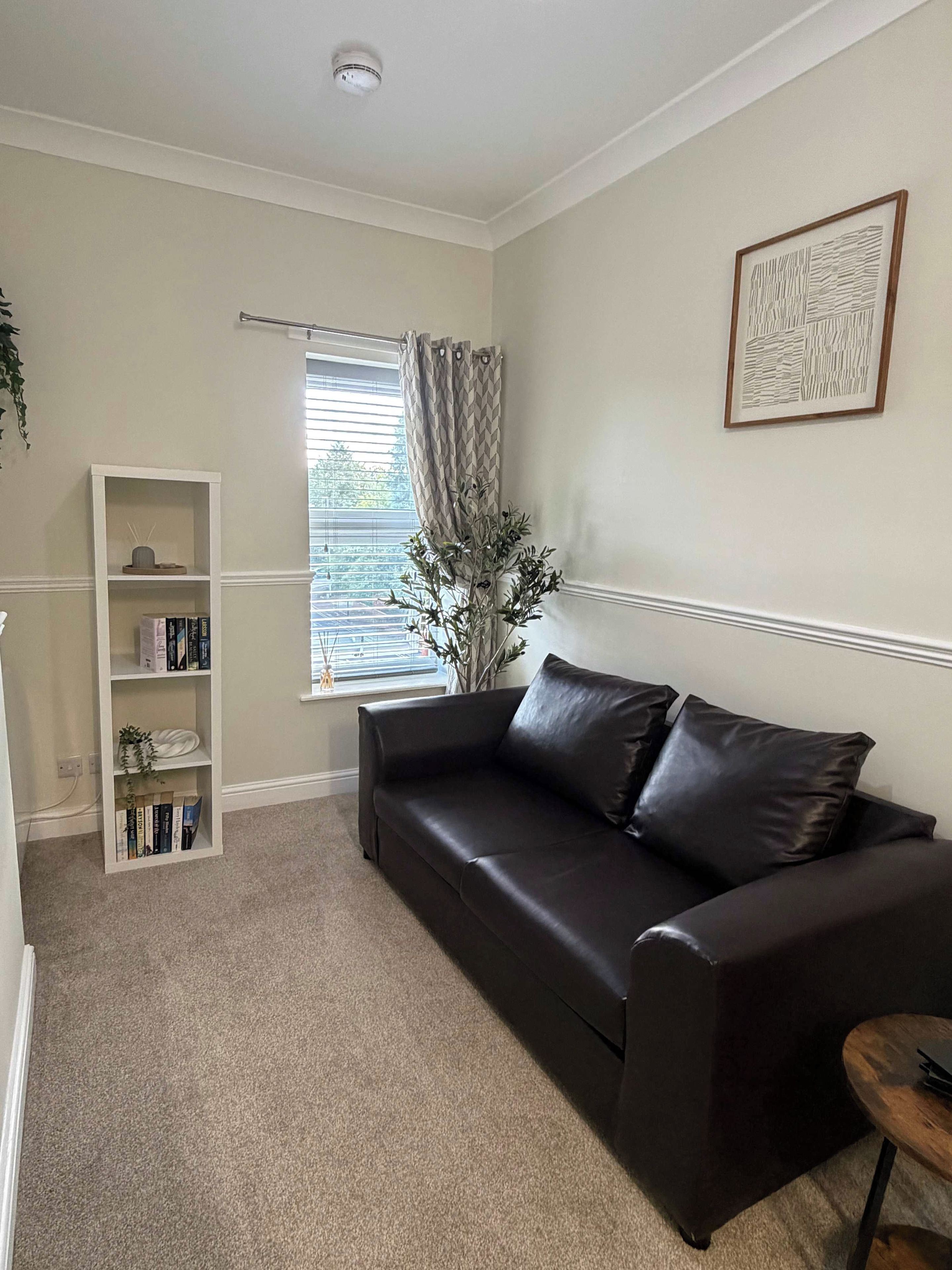 A living room with a dark brown leather sofa, a white bookshelf, a window with curtains, and a framed artwork on the wall.
