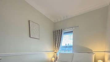 Een netjes opgemaakt bed met witte lakens en handdoeken, een bedlampje, een raam met gordijnen en een aan de muur hangend schilderij.