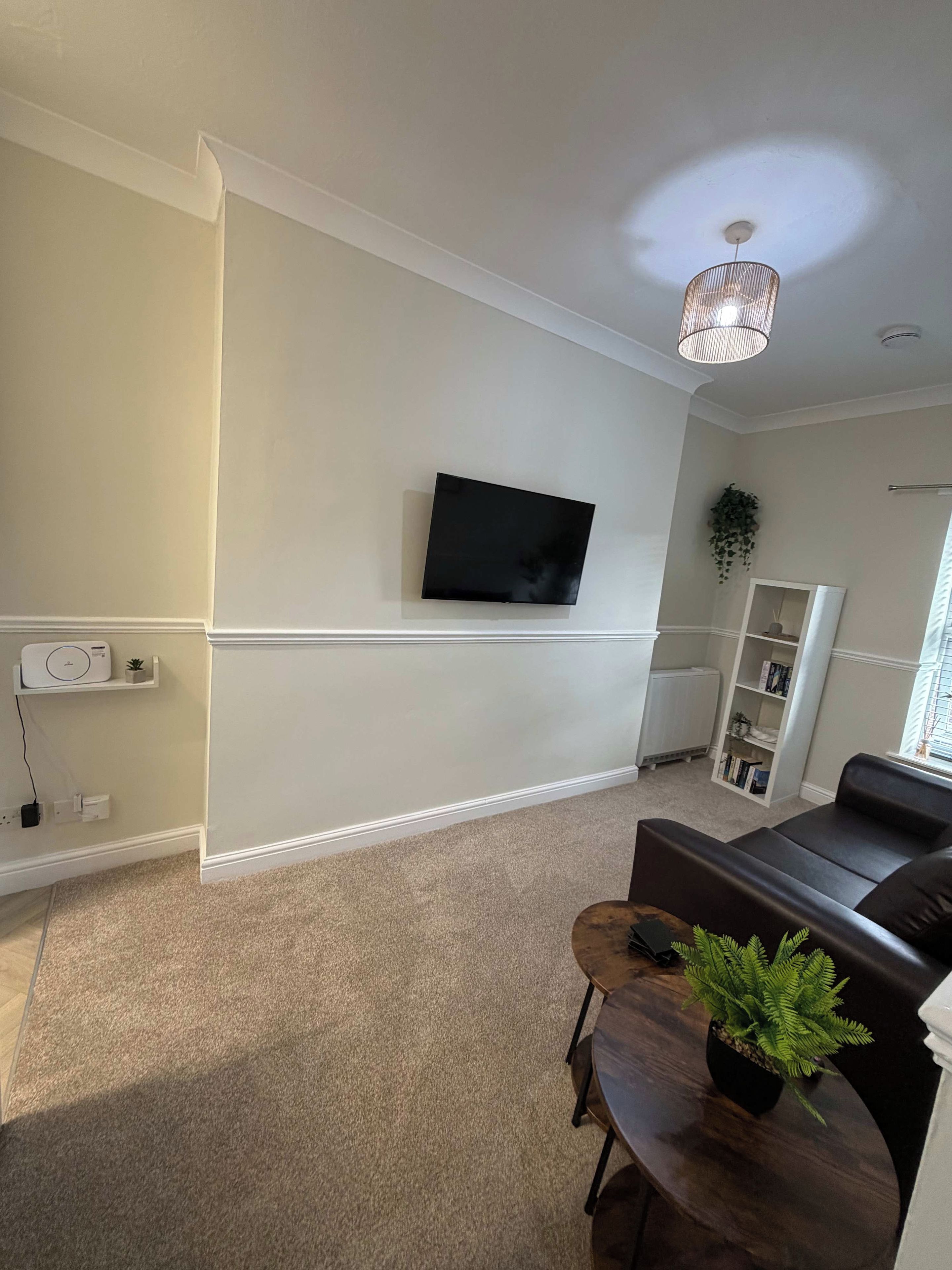 A living room with a flat-screen TV mounted on the wall, a dark leather sofa, a small wooden coffee table with a plant, and a white radiator.