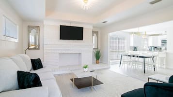 A modern living room with a white brick fireplace, a flat-screen TV mounted above it, a grey sofa with black cushions, a dark coffee table, and a view into the kitchen area.