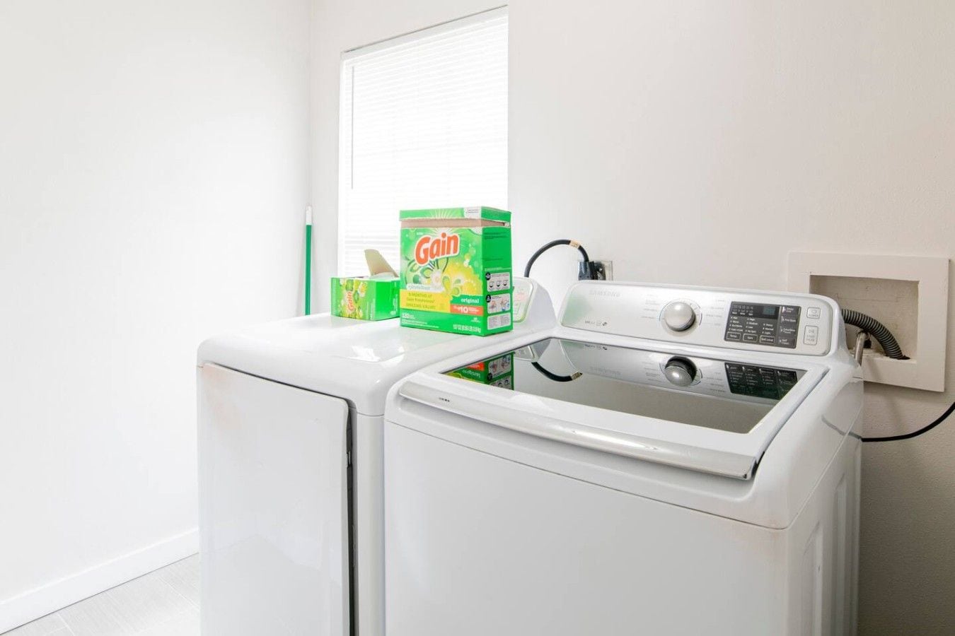 A front-loading washing machine with a control panel, a box of Gain detergent, and a power cord plugged into a wall socket.