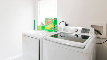 A front-loading washing machine with a control panel, a box of Gain detergent, and a power cord plugged into a wall socket.