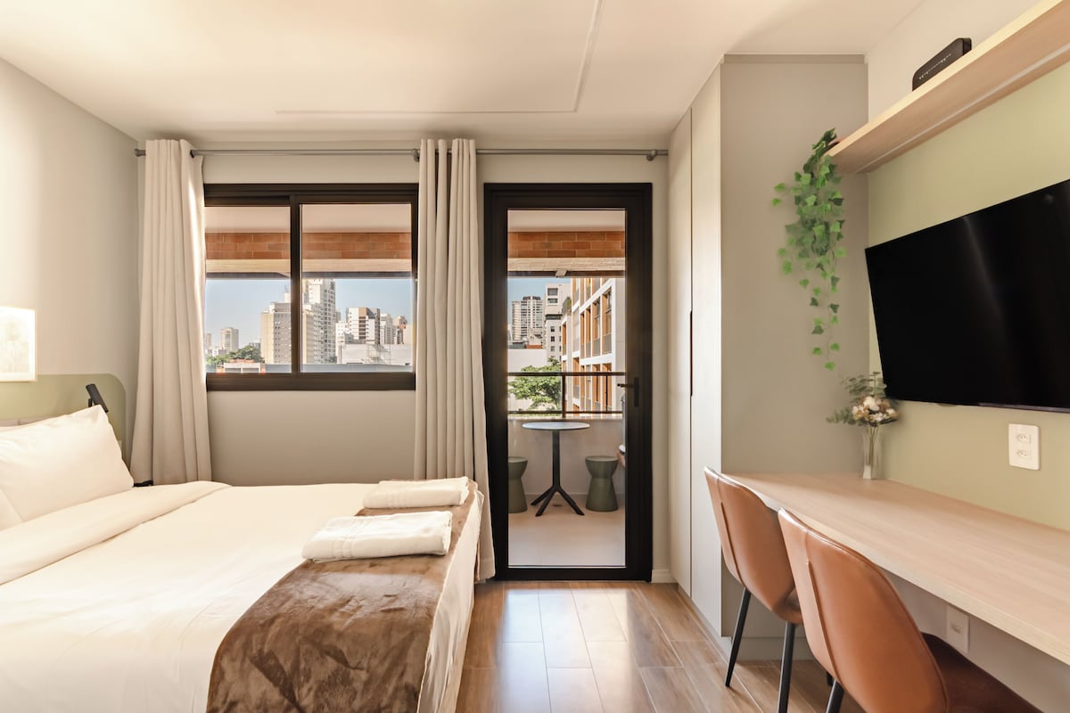 Een hotelkamer met een bed, bureau, stoel, televisie en een balkon met uitzicht op de stad.