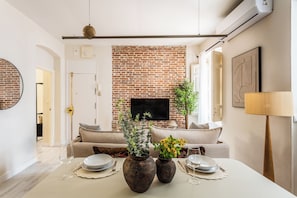 A living room with a brick wall, a sofa, a coffee table with decorative vases, and a television mounted on the wall. - A two-bedroom ground-floor home in Chamberí, where a private patio fills every room with light, and an exposed-brick dining area sets the scene for long Madrid evenings. (MADRID)