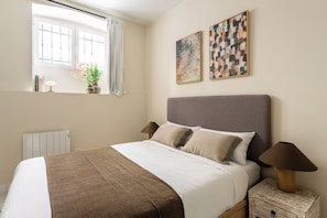 A neatly made bed with a grey headboard, two bedside lamps, and a window with a view of a bridge. - A two-bedroom ground-floor home in Chamberí, where a private patio fills every room with light, and an exposed-brick dining area sets the scene for long Madrid evenings. (MADRID)