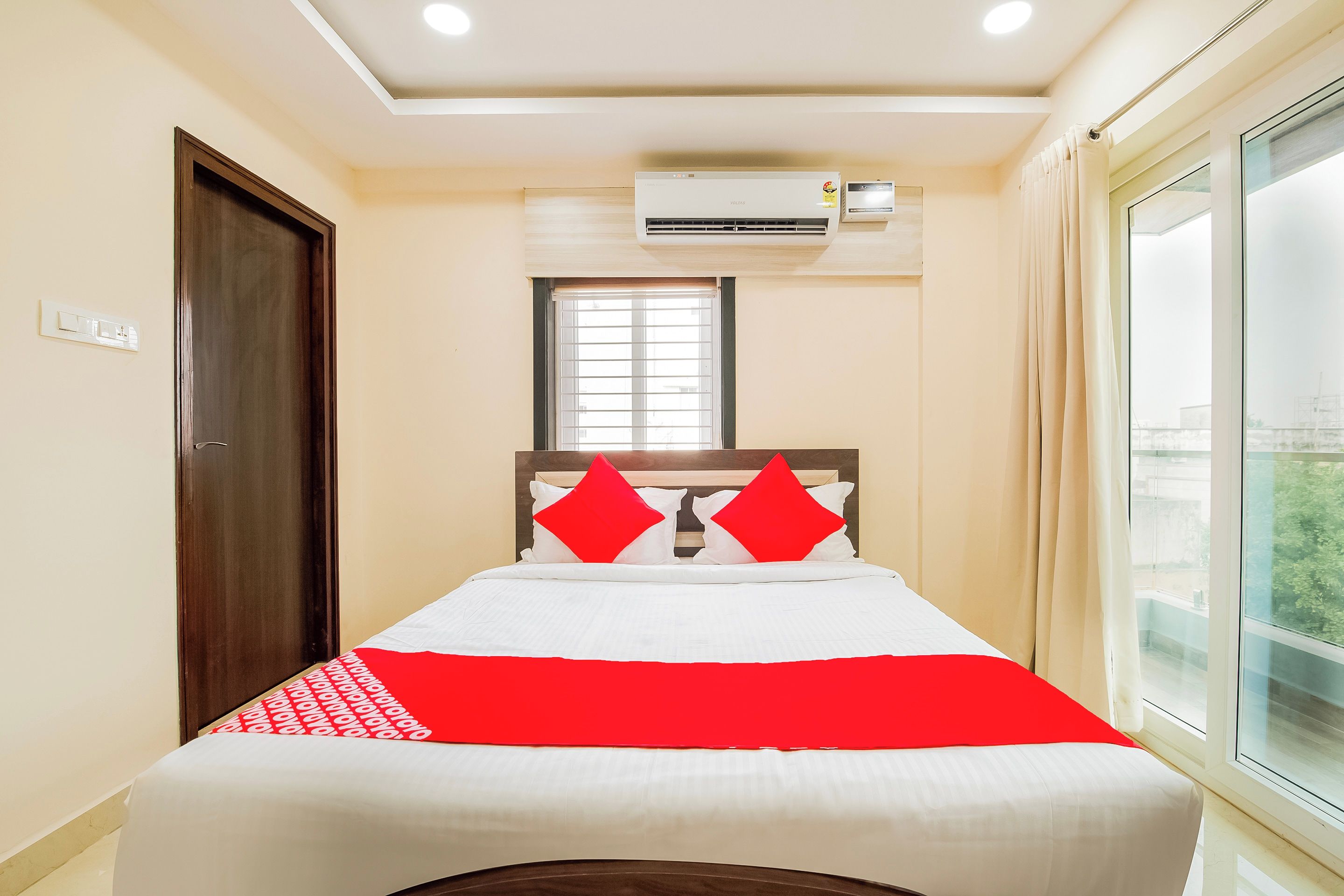 A hotel room with a bed, a window, an air conditioning unit, and a door.