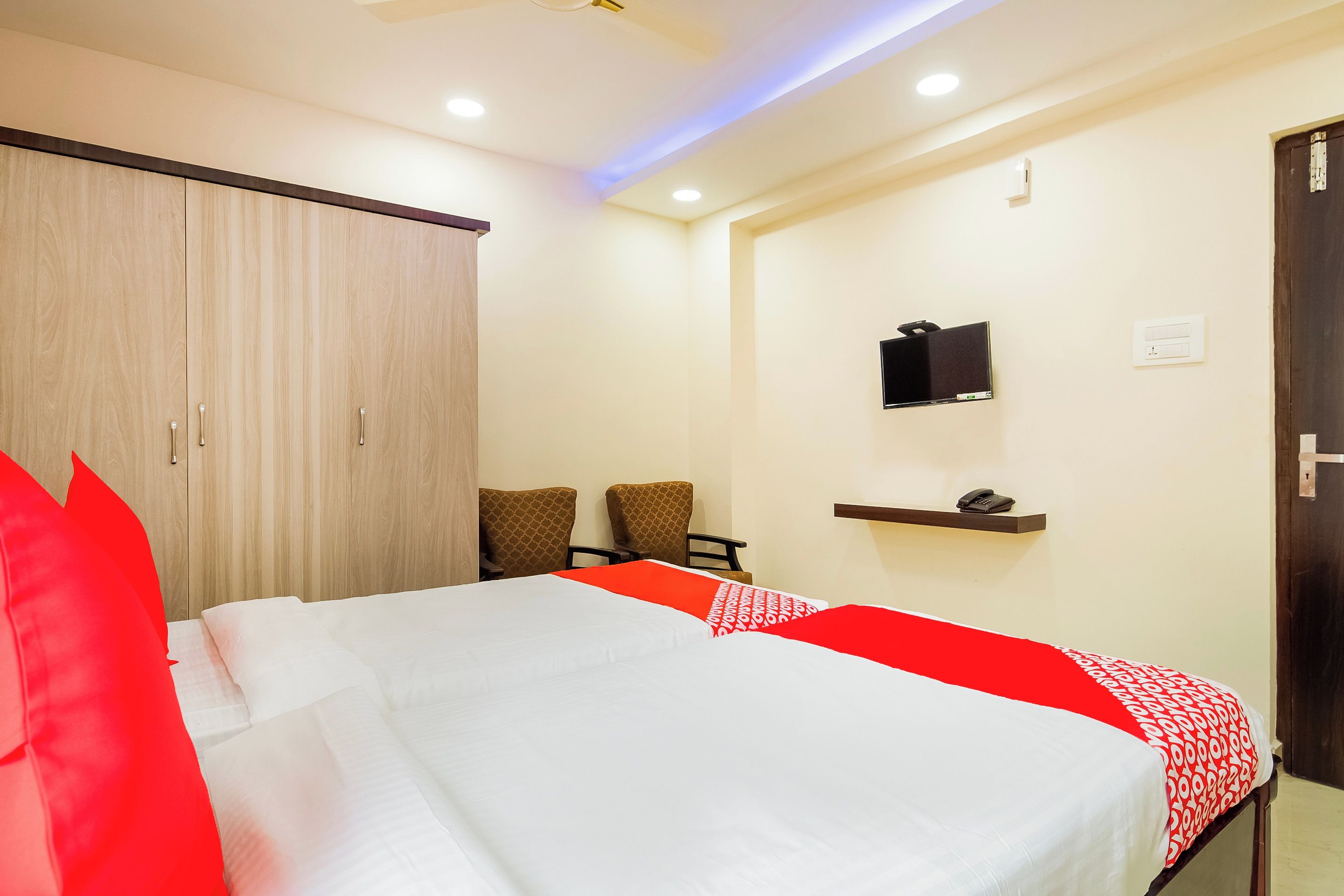 A hotel room with a bed, two chairs, a wardrobe, a shelf with a phone, and a TV on the wall.