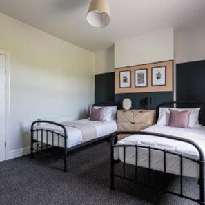 A bedroom with two beds, a wardrobe, and a wall-mounted shelf with framed pictures. - Centralbishopauckland - House - Stay6 - Nearcastle (Bishop Auckland)